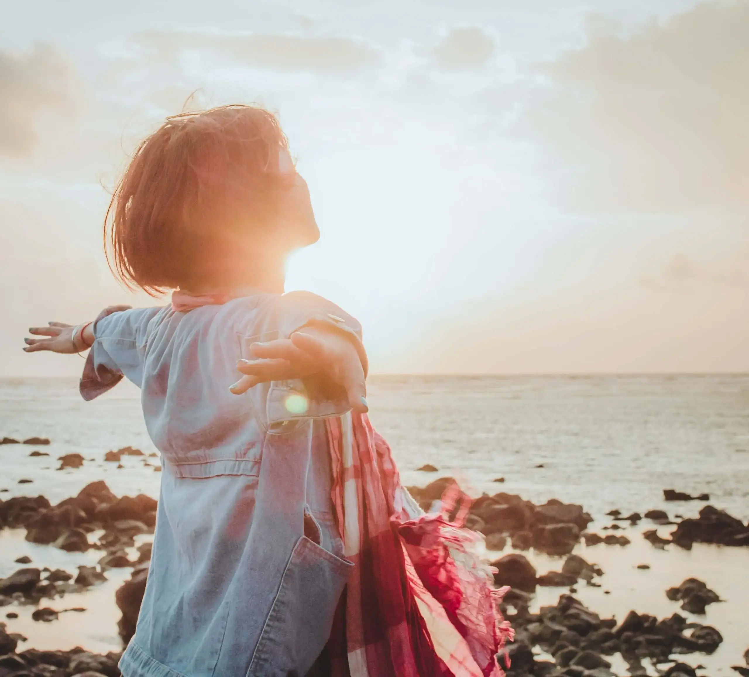 A woman standing by the ocean with arms outstretched, bathed in warm sunrise light, symbolizing freedom and confidence after overcoming fear