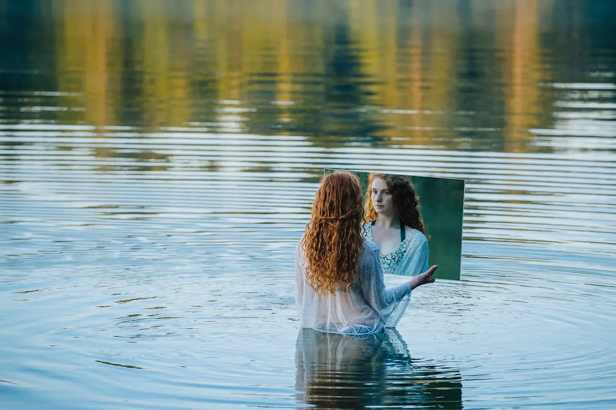 Person holding a mirror over a calm lake, reflecting their face; symbolizes introspection, self-reflection, and exploring inner self-image.