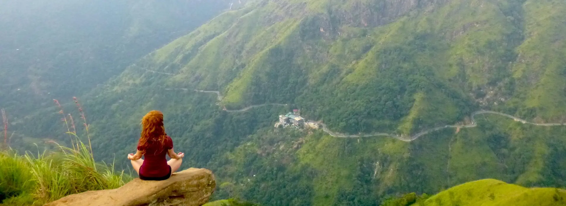 Woman sitting at the tip of a cliff, facing mountains, meditating in a serene landscape.